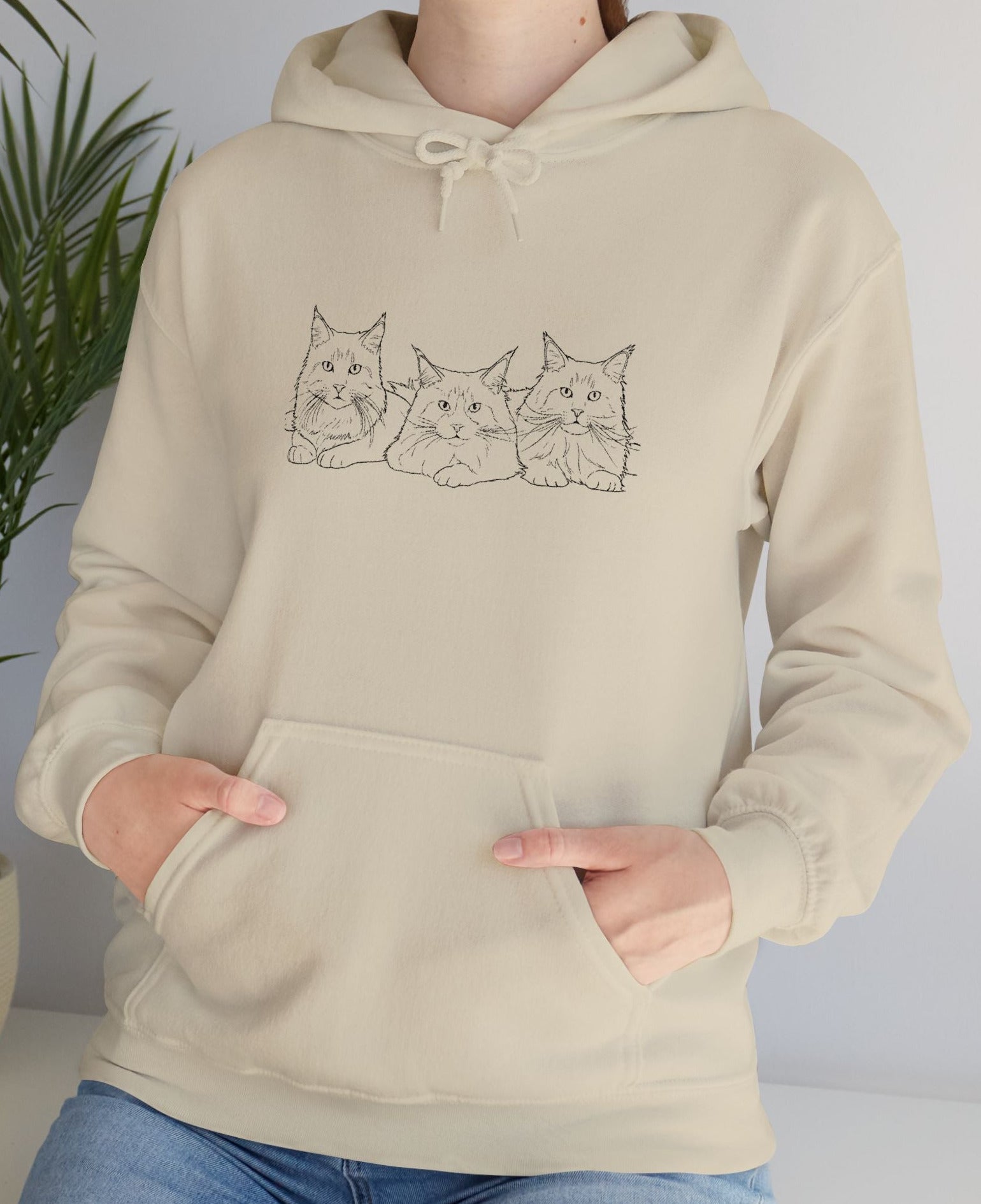 Person wearing a beige hoodie with cat design in front of a plant