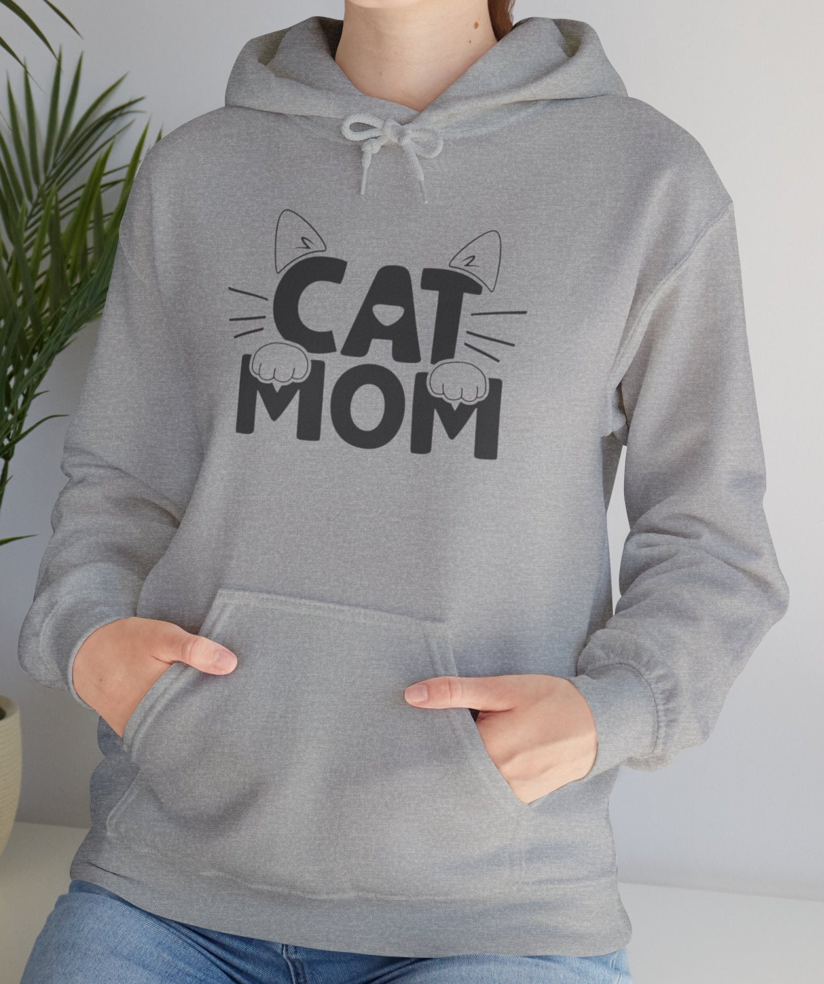Person wearing a gray hoodie with 'CAT MOM' text and cat graphics, standing indoors with a plant in the background.