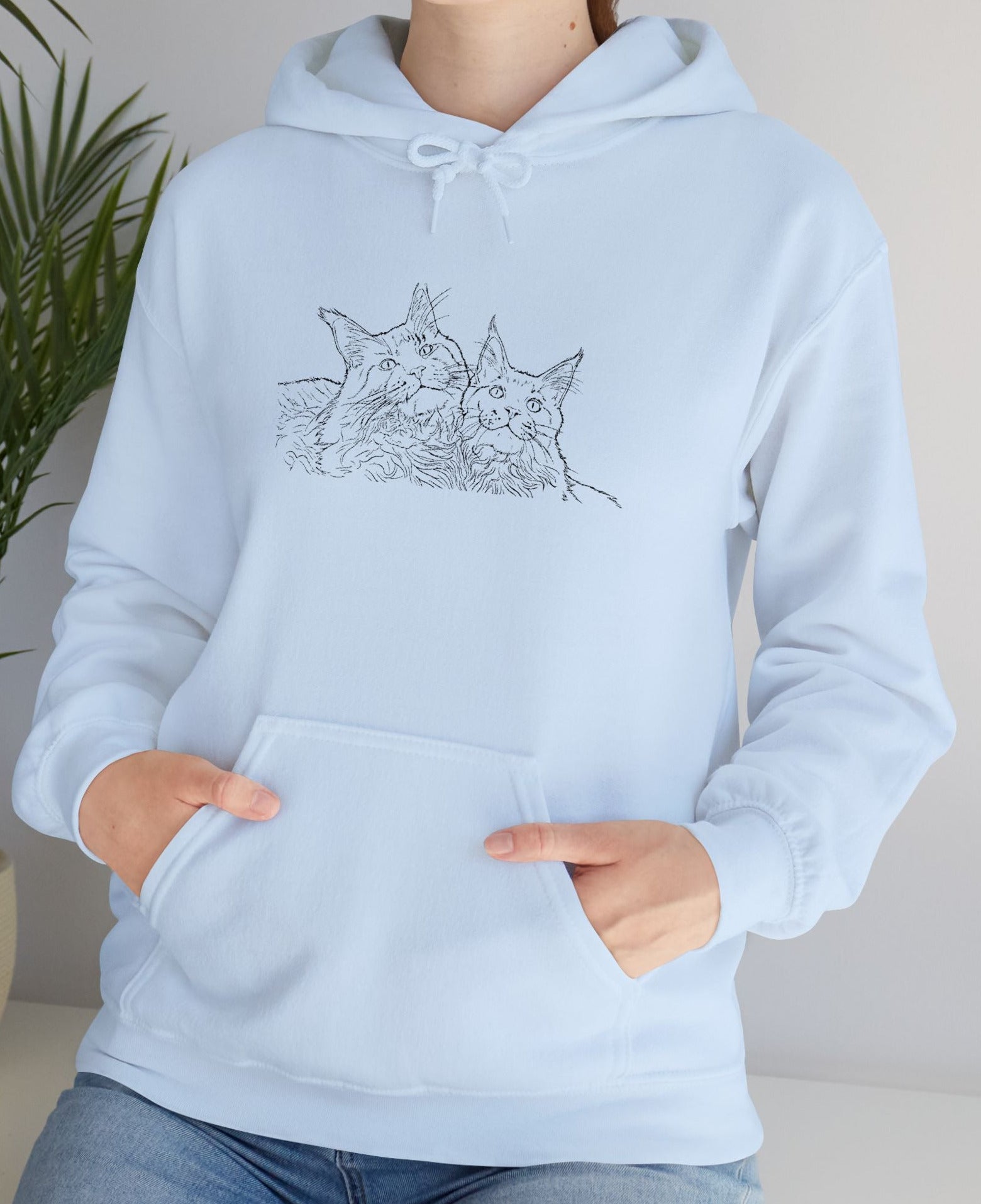 Person wearing a light blue hoodie with a cat design, standing indoors next to a plant.