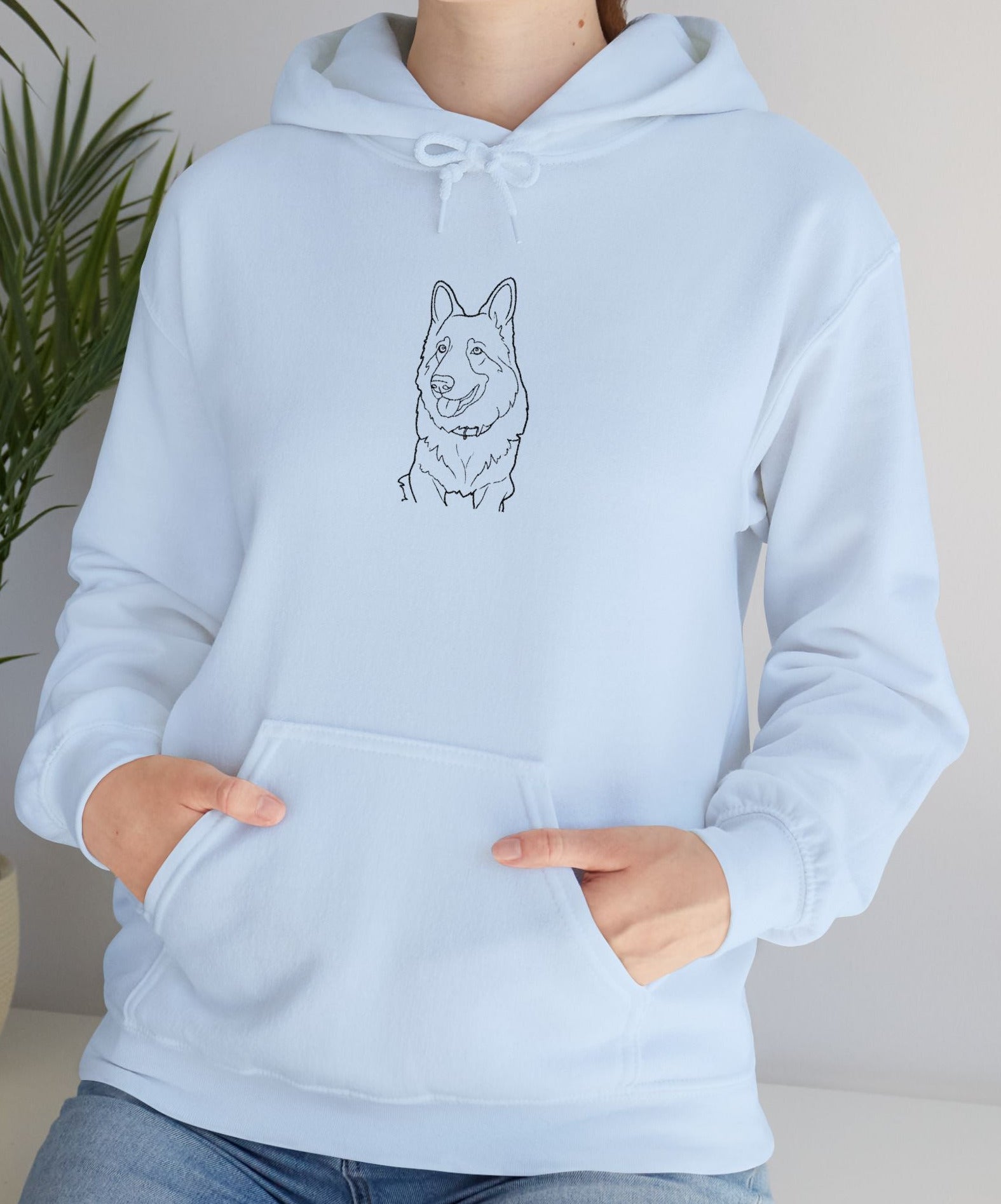 Person wearing a light blue hoodie with a dog illustration, standing indoors next to a plant.