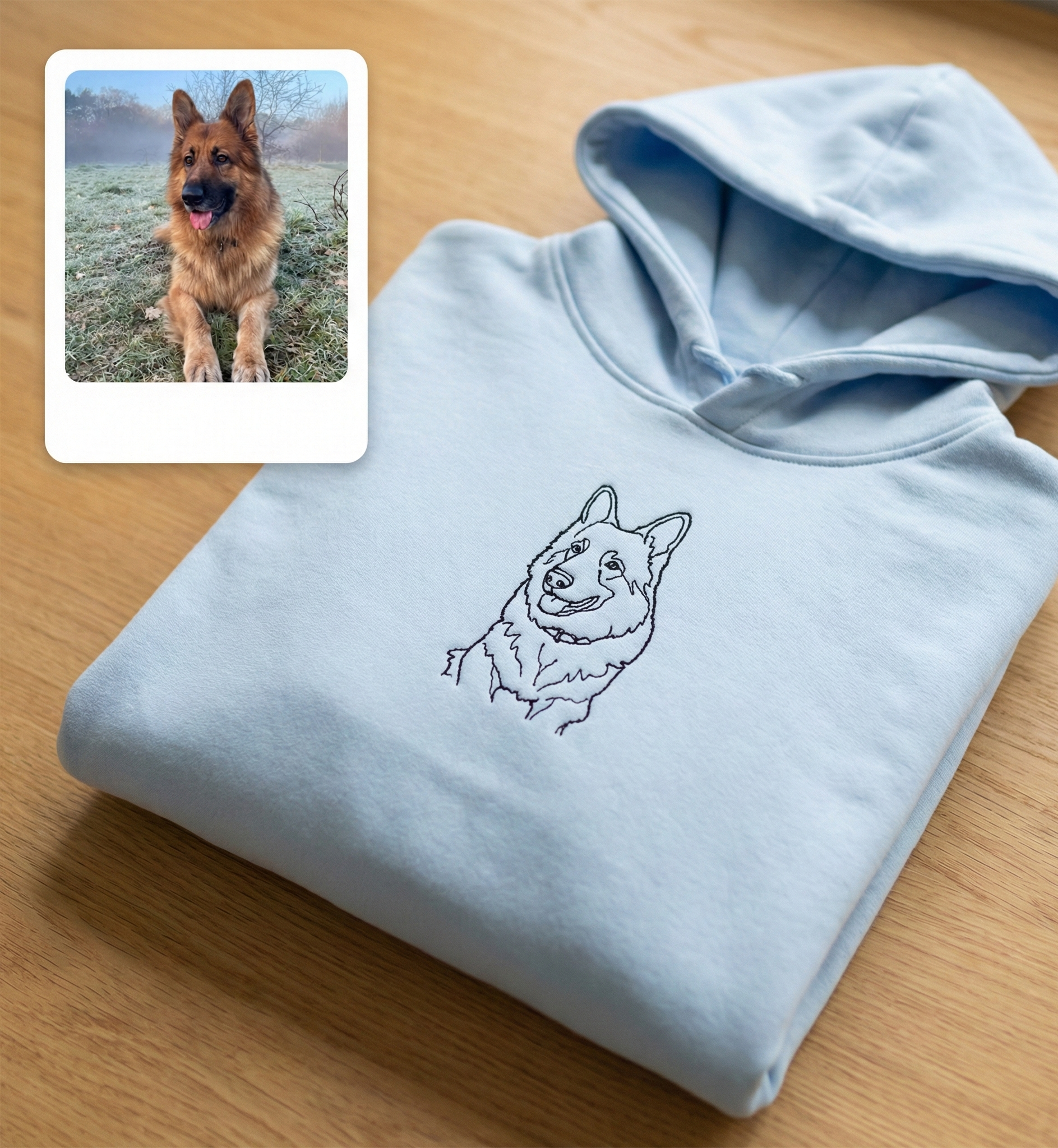 Folded light blue hoodie with a dog illustration on a wooden surface, with a small inset photo of a real dog.