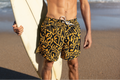 Man holding a surfboard on a beach wearing patterned shorts.